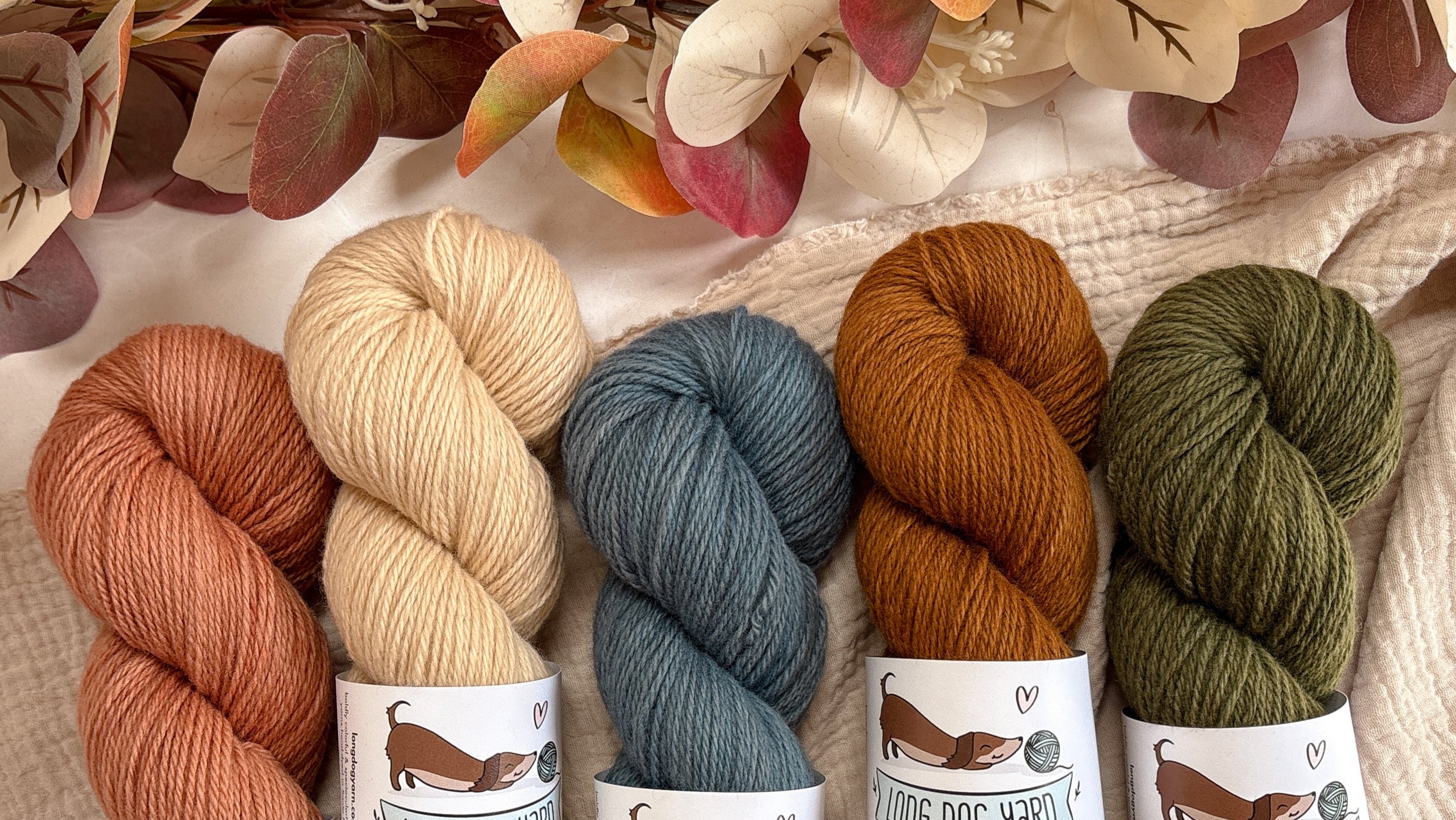 Five skeins of yarn in earthy tones with labels on a textured fabric background.